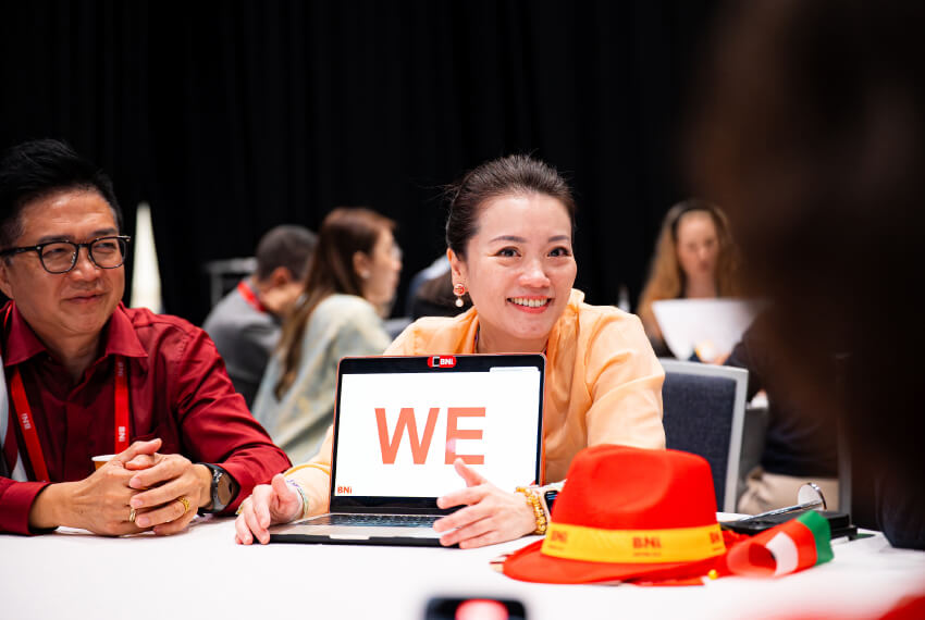 BNI members seated at a table during a meeting, with a laptop displaying the word “WE.”